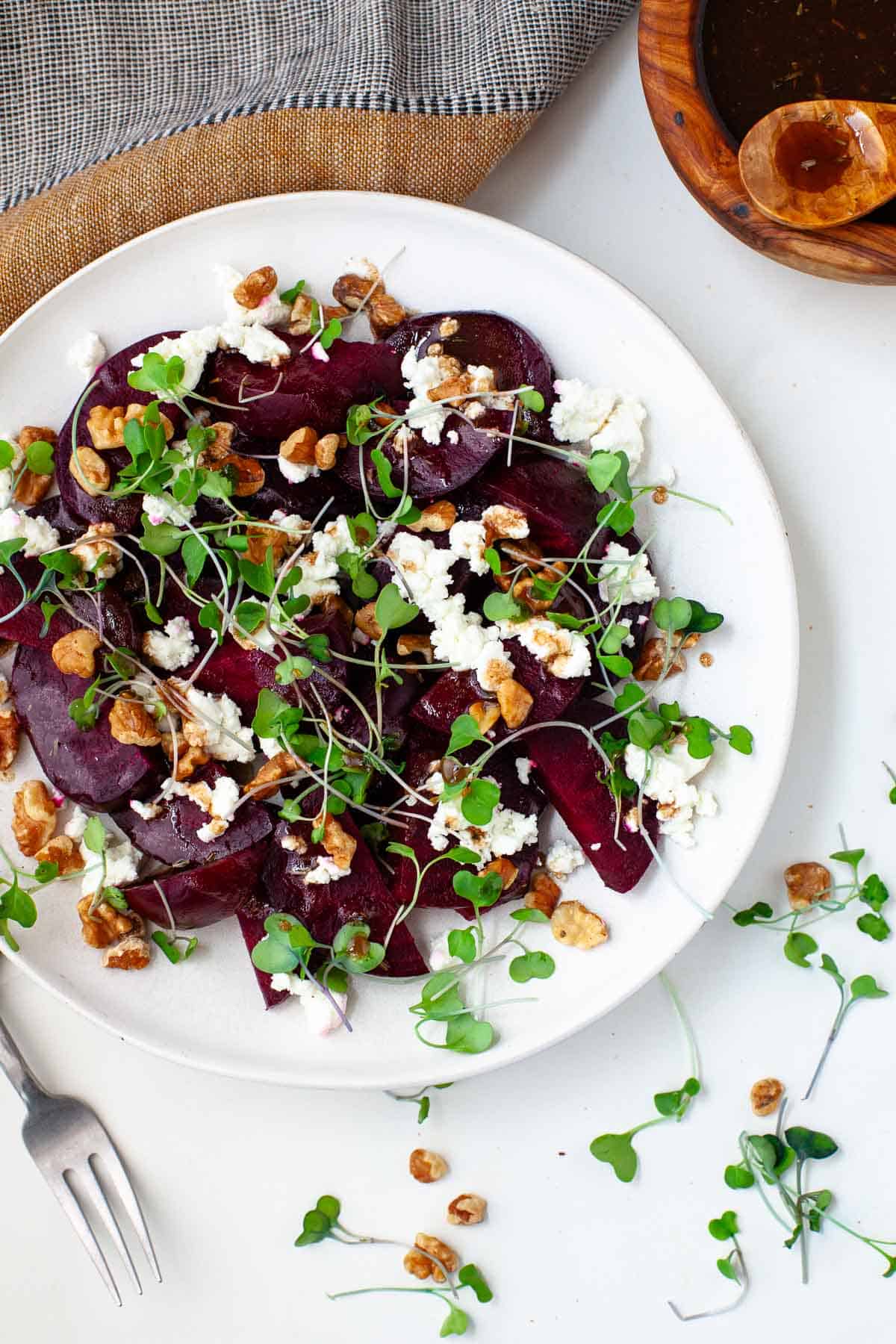 Beetroot salad with feta on white plate beside kitchen towel and small wooden bowl of balsamic vinaigrette.