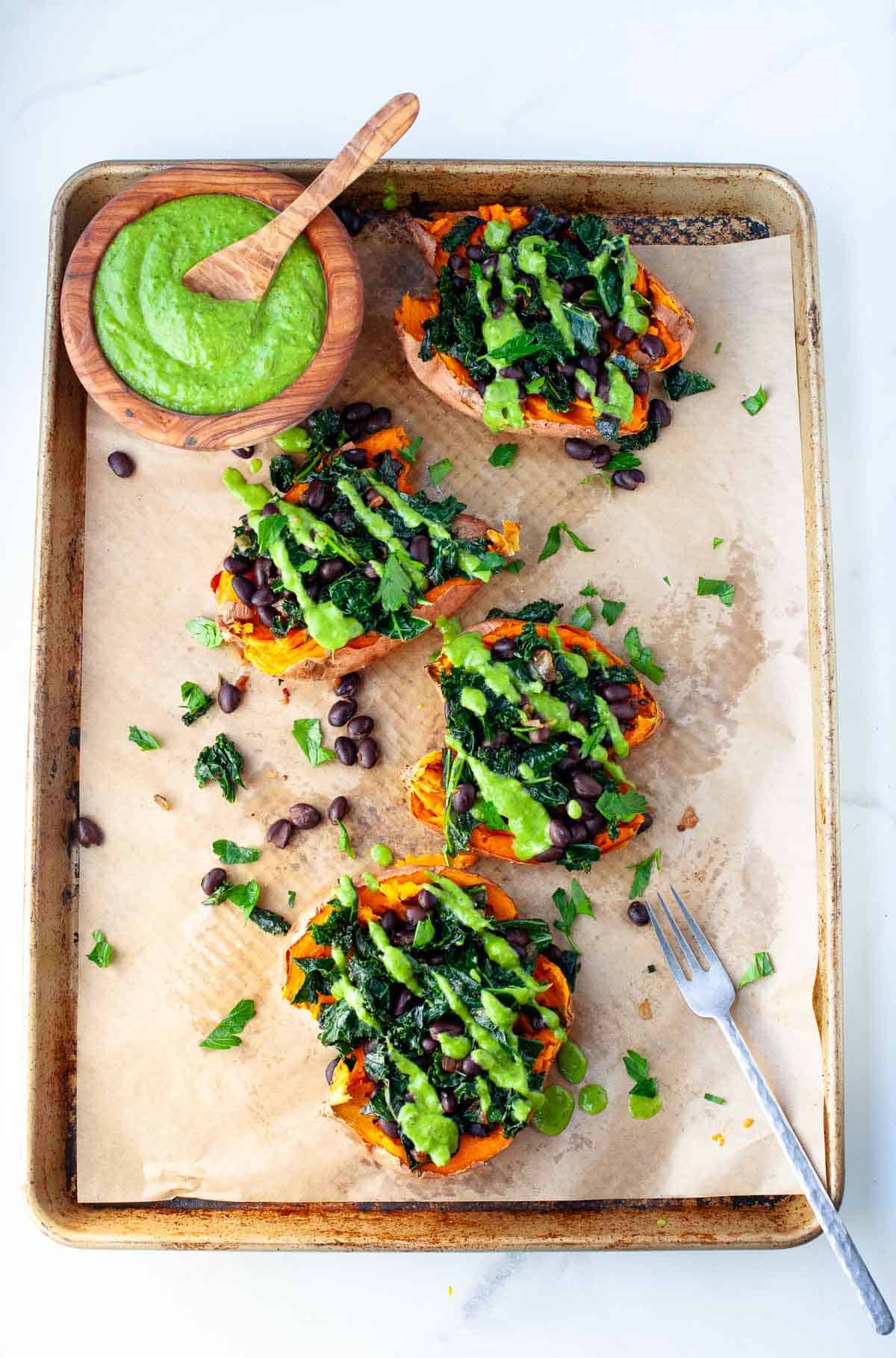 4 vegan loaded sweet potatoes drizzled with green goddess dressing and topped with fresh parsley with a side of dressing in a wooden bowl with spoon.