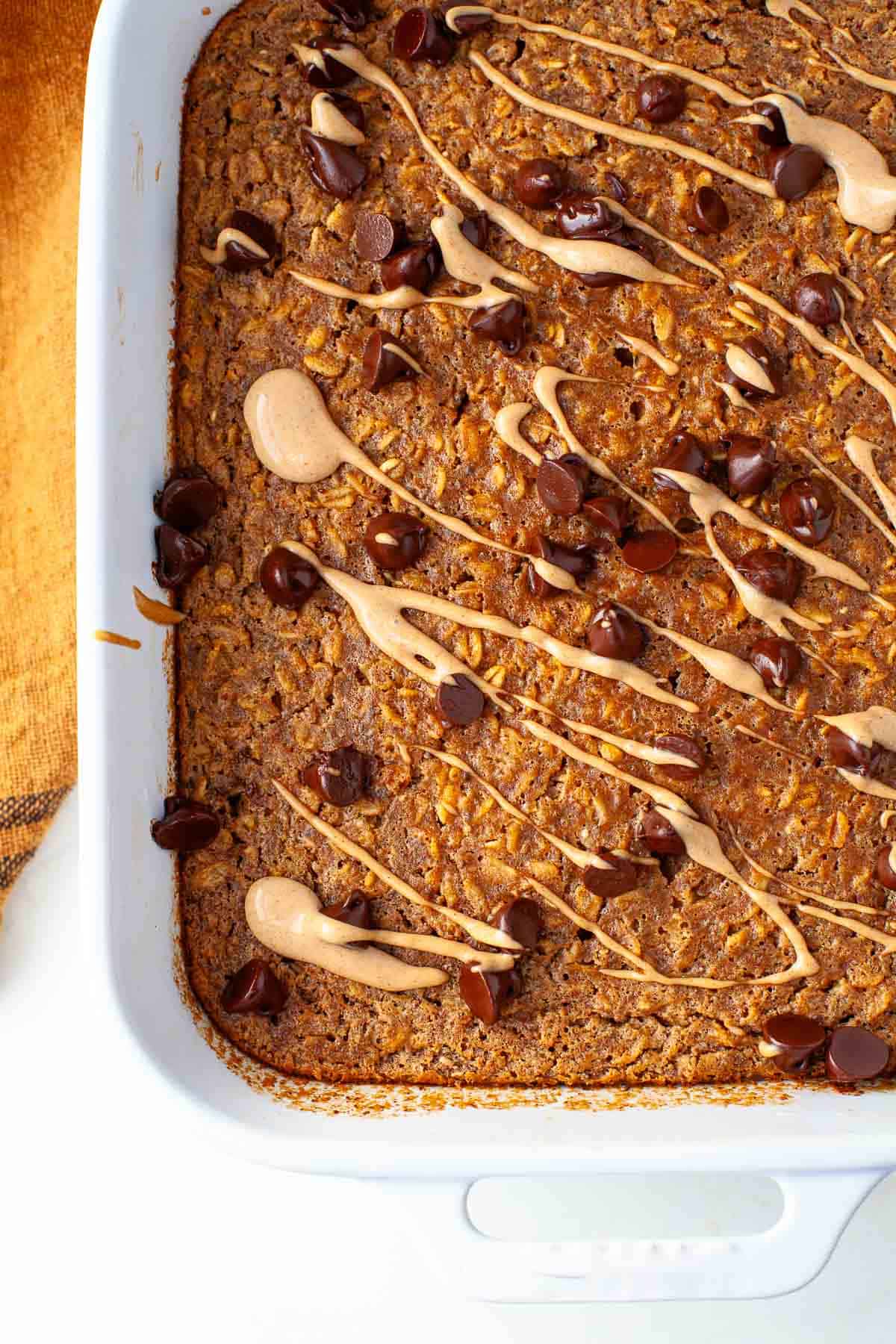 Peanut Butter Baked Oatmeal in white baking dish topped with chocolate chips and drizzled peanut butter.